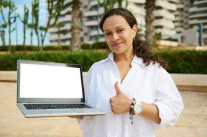 Confident woman real estate agent hold laptop with white mockup screen, shows thumb up at camera against modern building photo