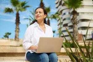 Portrait of a confident successful young woman working online sitting on a stone bench against modern buildings backdrop photo