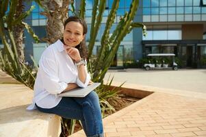 Beautiful woman in casual clothes, sitting on a stone bench with laptop on her knees, smiling cutely looking at camera photo