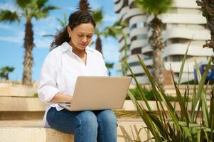 Confident multitasking business woman sitting on a stone bench with laptop on her knees, typing text, online working photo