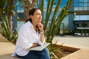 Beautiful woman in casual clothes, sitting on a stone bench with laptop on her knees, smiling and thoughtfully looking away photo