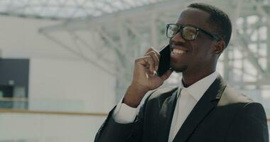 Smiling African American businessman in suit speaking on mobile phone discussing cooperation in sunlit office building lobby. Communication and modern employee concept. video