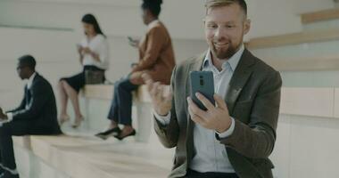 Joyful businessman making online video call with smartphone sitting outside office center in city enjoying work break. People and communication concept.