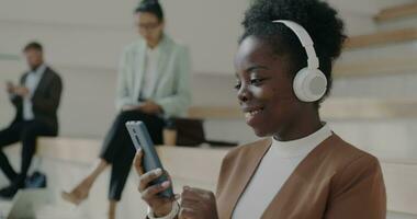 African American businesswoman listening to music with headphones using smartphone relaxing on steps of office center. Entertainment and work break concept. video