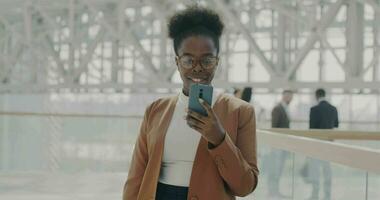 långsam rörelse porträtt av framgångsrik ung affärskvinna använder sig av smartphone stående i kontor byggnad lobby njuter företags- kommunikation video