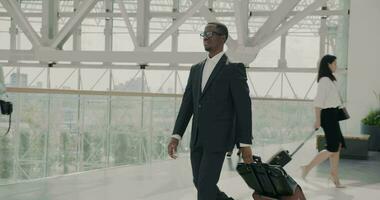 dolly tiro di elegante africano americano uomo d'affari a piedi nel aeroporto con valigia mentre persone con borse in movimento intorno a. moderno città visibile nel panoramico finestre. video