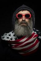 An elderly man with a gray beard in a hood and a scarf with a pattern of the American flag on a black background. photo