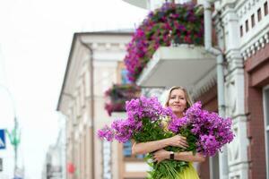 un hermosa mayor mujer con un ramo de flores de flores camina alrededor el ciudad. años modelo rubio con azul ojos es contento. un mujer de cincuenta años camina en el verano, teniendo un bueno tiempo. foto