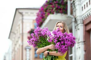 un hermosa mayor mujer con un ramo de flores de flores camina alrededor el ciudad. años modelo rubio con azul ojos es contento. un mujer de cincuenta años camina en el verano, teniendo un bueno tiempo. foto