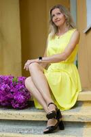 A beautiful elderly woman with a bouquet of flowers is sitting on the stairs. Age model blonde with blue eyes is happy. A woman of fifty years walks in the summer, having a good time. photo