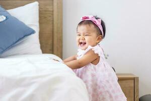 Asian baby toddler in cute pink dress is smiling while start practice walking and balancing herself at the bed with happiness for healthy kid and adorable girl portrait usage photo
