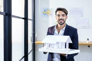 Young caucasian architect is holding house model inside working office with floor plan layout for real estate and property development project in eco friendly design and security solution concept photo