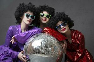 A group of disco girls in wigs with a disco ball and colorful costumes pose against a gray background. Disco party. photo