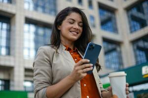 Young smiling brunette woman holding a smartphone and a cup of coffee looking at device screen against the background of an office building. copy space photo
