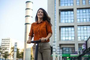 Next generation commute to work using e-scooters by female business persons. Young beautiful woman in a blue jacket smiles and rides an electric scooter to work along office buildings photo