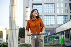 Next generation commute to work using e-scooters by female business persons. Young beautiful woman in a blue jacket smiles and rides an electric scooter to work along office buildings photo