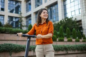 Young beautiful woman smiles and rides an electric scooter to work along office buildings. Ecological transportation concept. photo