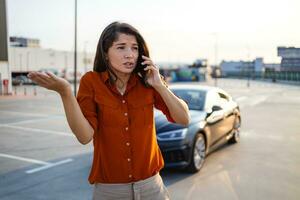 nervioso joven mujer en pie por abierto coche capucha y vocación a auto servicio, tiene roto a autopista en un camino a campo. hembra conducir teniendo teléfono conversacion con amigos, preguntando para ayuda con auto foto