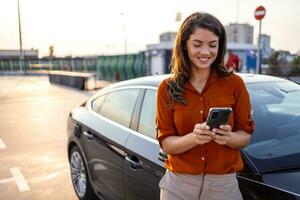 Woman using mobile phone, communication or online application, standing near car on city street or parking, outdoors. Car sharing, rental service or taxi app. photo