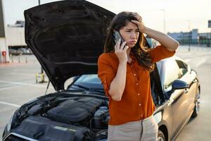 nervioso joven mujer en pie por abierto coche capucha y vocación a auto servicio, tiene roto a autopista en un camino a campo. hembra conducir teniendo teléfono conversacion con amigos, preguntando para ayuda con auto foto