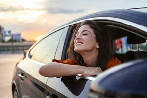 alegre joven hembra sentado en brillante coche en pasajero asiento y propensión fuera abierto ventana mientras disfrutando el paseo foto