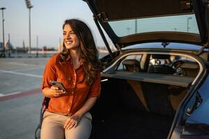 Young woman sitting in the car trunk with a smart phone. Beautiful female laughing in car with mobile phone. photo