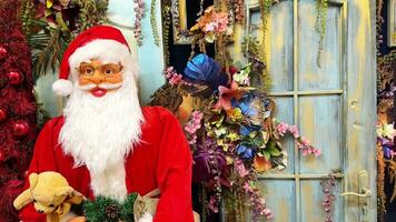 Papa Noel claus con un suave juguete en su manos soportes en frente de el puerta de un Tienda con Navidad decoraciones y da la bienvenida huéspedes. gracioso juguete Papa Noel claus en el escaparate. de cerca. video