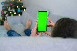 Closeup shot of man  hand using mobile phone with Green screen against the backdrop of a Christmas tree. Finger swiping up and down in green screen of the mobile. Green screen mockup template . photo