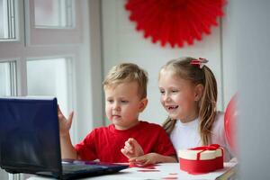 Happy kids waving, via laptop celebrating February 14 during quarantine, self-isolation and family values, online - Valentine's Day. Children's congratulations to friends via the Internet photo