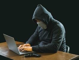 Portrait hacker spy man one person in black hoodie sitting on table looking computer laptop used login password attack security to data digital internet network system night dark background copy space photo