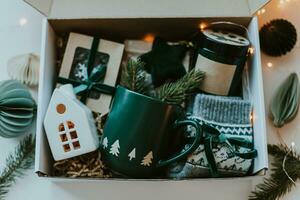Christmas care package gift box. Paper box with cozy mug, candles, socks, candies and white ceramic house. Christmas and Holiday Gift Ideas photo