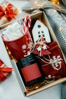 Christmas box with presents in red colors. Candle in dark jar, mug with marshmallow, white ceramic house, red socks and festive decoration. Top view photo