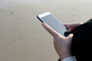 Closed up hand holding smartphone and touch screen for online with internet on the beach and seaside for business trip. Vacation and relaxation with technology on holiday concept photo