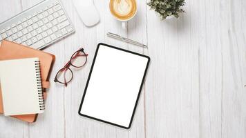 White wooden desk with blank screen tablet, keyboard, mouse, eyeglass, notebook, pen and cup of coffee, Top view flat lay with copy space. photo