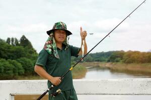 An Asian man holding a fishing rod and his catch stands on a bridge over a river in the village where he is a regular fisherman on holiday. And it's a fun and relaxing outdoor activity. photo