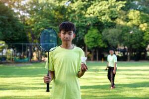 An Asian boy holds a badminton racket and a white shuttlecock while playing badminton with friends on the park lawn in the evening after returning from school. photo