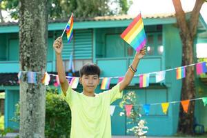Asian boy holds a rainbow flag in front of a house decorated with rainbow flags during Pride Month to show LGBT pride and identity. Soft and selective focus. photo