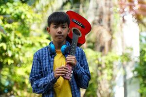 Asian boy carrying a classical guitar on his shoulder to play with friends at a summer camp at a national park. Soft and selective focus. photo