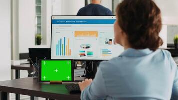 Office worker checks greenscreen display on tablet, working on business operations with isolated copyspace template and analyzing data on computer at desk. Having gadget with mockup. video