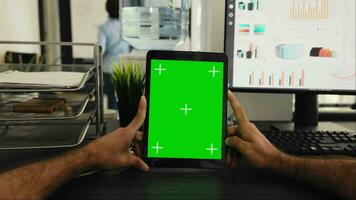 POV of businessman holding tablet with greenscreen template, looking at chromakey display and sitting coworking space desk. Person examining isolated mockup copyspace on gadget screen. video