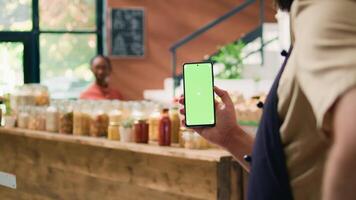 lokaal verkoper houdt smartphone app met groene scherm Scherm, presenteren copyspace technologie Aan mobiel apparaat. jong Mens tonen geïsoleerd mockup scherm Bij kruidenier op te slaan uitchecken. video