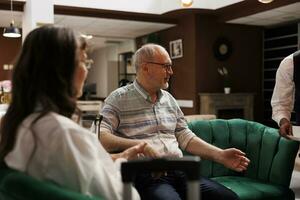 Elderly couple on comfortable sofas receiving assistance from helpful resort employee. Retired senior man receiving cup of coffee from african american waiter while talking to his wife. photo