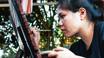 de cerca de asiático mujer pintor creando Arte utilizar un Cepillo de pintura a dibujar letras diseños en un de madera café tienda signo. al aire libre actividades, personas haciendo actividades. foto