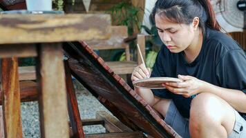 asiático mujer pintor creando Arte utilizar un Cepillo de pintura a dibujar letras diseños en un de madera café tienda signo. al aire libre actividades, personas haciendo actividades. foto
