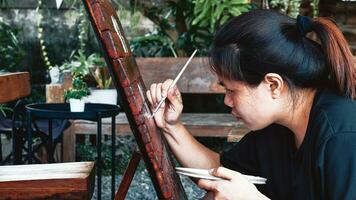 asiático mujer pintor creando Arte utilizar un Cepillo de pintura a dibujar letras diseños en un de madera café tienda signo. al aire libre actividades, personas haciendo actividades. foto