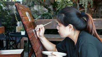 asiático mujer pintor creando Arte utilizar un Cepillo de pintura a dibujar letras diseños en un de madera café tienda firmar.al aire libre actividades, personas haciendo actividades. foto
