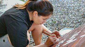 asiático mujer pintor creando Arte utilizar un Cepillo de pintura a dibujar letras diseños en un de madera café tienda signo. al aire libre actividades, personas haciendo actividades. foto