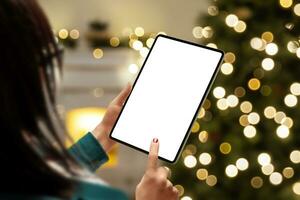 Woman with tablet mockup by a Christmas tree, offering an isolated display screen for app or web page presentation. Ideal for showcasing apps or web designs in a festive setting photo