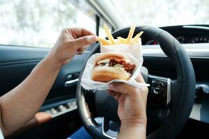 Asian woman driver hold and eat hamburger and french fries in car, dangerous and risk an accident. photo