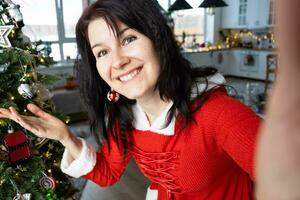 Selfie portrait of a woman in a red Santa dress in a home interior with a Christmas tree and New Year decor. Preparation for the holidays, party photo